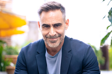 A middle-aged businessman in a stylish suit smiling confidently against a backdrop of modern office buildings. His professional and approachable demeanor reflects success and leadership. 