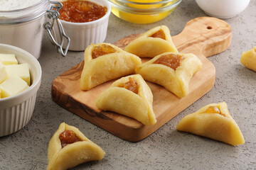 Raw sugar cookies in a triangular shape with filling on a wooden cutting board, preparation for the traditional Jewish holiday Purim day. A group of ingredients on the background. side view