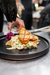 A close-up of a chef plating grilled chicken on a black plate, accompanied by creamy sauce and garnished with purple cabbage. The background features a busy kitchen setting.