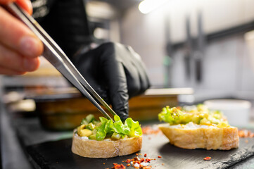 A chef's hand in a black glove uses tweezers to carefully place fresh greens on a gourmet appetizer. The dish is presented on a slate surface, showcasing attention to detail in culinary presentation.