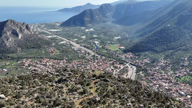 Aerial video of the Greek flag on a cliff overlooking Leonidio town with red cliffs and stone houses, Peloponnese, Greece.