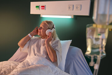 Elderly woman in hospital gown sitting in bed in the evening and listening to relaxing music in...