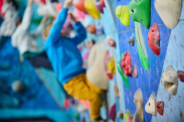 Fototapeta premium Indoor climbing gym with a focus on colorful holds. In the blurred background, a young boy in a blue hoodie and yellow pants is climbing, adding depth and action to the scene