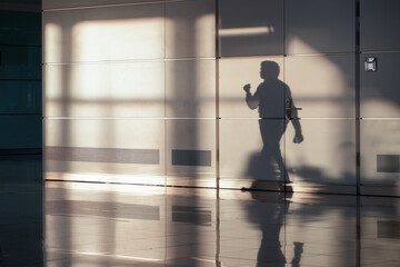 A lone figure's shadow stretches across a textured airport wall, creating an abstract light pattern. This monochrome image encapsulates a moment of anonymity and transition in a contemporary space