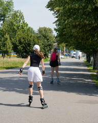 Fototapeta premium Two women roller skating on a tree-lined road path during a sunny summer day. Both skaters are equipped with helmets and knee pads, embracing fitness and outdoor recreation in a picturesque setting
