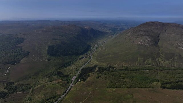 Barnesmore Gap, County Donegal, June 2023. Drone tracking left reveals road connecting towns crossing through forest and grassy boglands below stunning mountain ranges and hill terrain, summer day