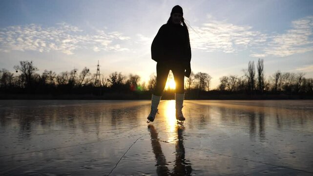 Silhouette of girl shod in figure skates sliding on pond during beautiful sunset. Sportswoman improving skills at lake on freeze day. Shining sun reflecting on surface of ice. Active leisure at winter