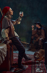 Two men, dressed like medieval prated raising glasses with rum, drinking again vintage green background in dimly-lit room. Concept of camaraderie, history, adventure, achievement