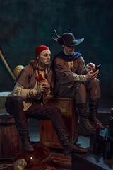 Fototapeta premium Young men, medieval pirates in vintage costumes sitting with smartphones, scrolling social media feed, discussing news against dark background in dimly-lit room. Concept of digital networking