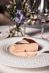 A slice of cheesecake is on a white plate with a vase of flowers in the backgrou