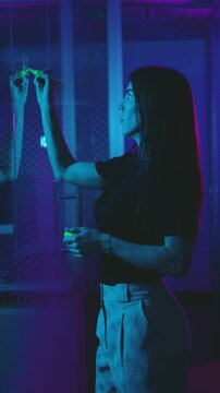 Real time side view of young female with long hair standing near reflecting glass wall and writing with phosphorescent marking pen make a difference words at night time
