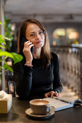 Woman on phone with coffee and notebook at cafe, woman in glasses talking on phone with coffee and notebook in cafe.