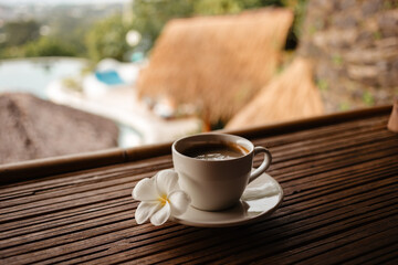 Morning hot Americano in a white mug with frangipani flower. Black coffee on a bamboo table. Cup of strong coffee in the morning on vacation