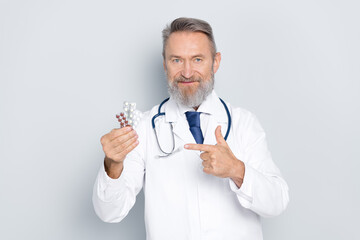 Confident senior doctor presenting medication while pointing, wearing a white coat, and standing against a neutral grey background.