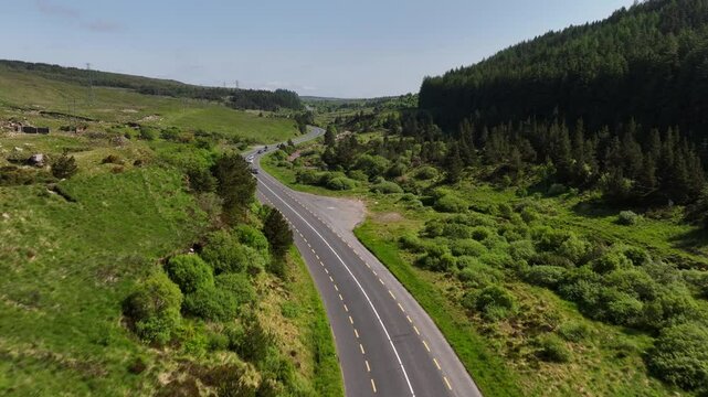 Barnesmore Gap, County Donegal, Ireland, June 2023. Drone pushes forwards as a cars navigate a winding road through the valley, crossing rocky rivers and bogland on a bright sunny day.