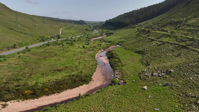 Barnesmore Gap, County Donegal, June 2023. Drone low approach to shallow waterway with rocks and sediment built up along riverbanks at base of steep slope next to road