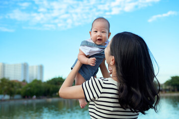 Joyful Mother Lifts Her Baby Near Tranquil Lake on a Sunny Day in a Vibrant Outdoor Setting