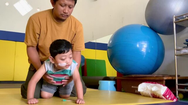 A father trains a toddler who lives with cerebral palsy and installs an NGT nasogastric tube in his nose to learn to crawl in the physiotherapy room.