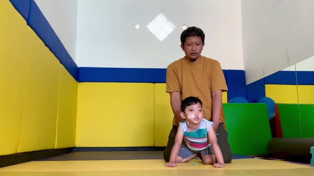 A father trains a toddler who lives with cerebral palsy and installs an NGT nasogastric tube in his nose to learn to crawl in the physiotherapy room.