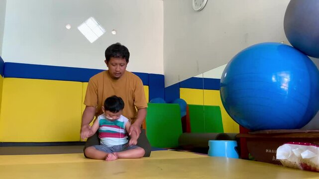 Male physiotherapists are helping and training boys who live with cerebral palsy to sit in colorful therapy room.
