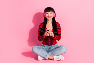 Young woman wearing headphones smiling while holding a phone against a pink background, dressed in casual striped outfit