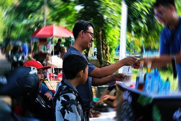A candid moment at a lively outdoor market, featuring a father and son near a colorful stall with various items on display, surrounded by greenery and bustling activity in the background.