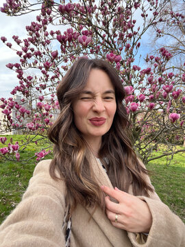 Caucasian woman with air touch hair coloring winks and taking a selfie near blooming magnolia tree
