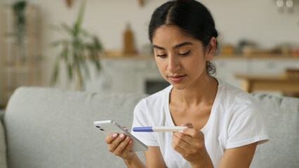 Worried anxiety puzzled confused Indian woman Arabian lady female girl holding positive result pregnancy test unwanted unplanned pregnant at home couch chatting mobile phone message health medicine