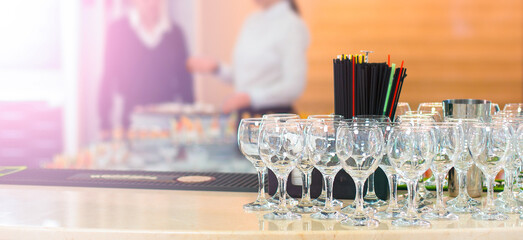 Bar counter with empty wine glasses, a container of straws, and blurred bartenders in the background.