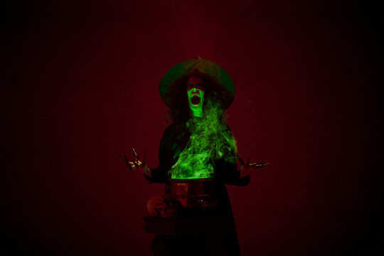 Shouting witch in black cloak and pointed hat making spell with bubbling cauldron and green mist rising around, in a dimly lit chamber. Concept of mystery, fantasy - Powered by Adobe