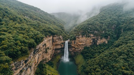 Naklejka premium Majestic waterfall cascades down steep cliffs, lush green mountains in foggy backdrop. Ideal for travel brochures