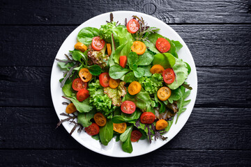 Mix of fresh organic juicy salad greens and red and yellow cherry tomatoes in white plate on black wooden table. Food photography