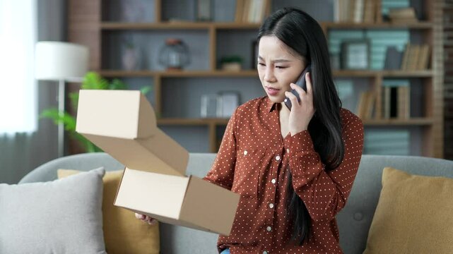 Unhappy young Asian woman arguing on the phone after receiving a parcel with wrong product at home. Angry female makes a bad review, she is dissatisfied with shopping in an online store. Damaged item