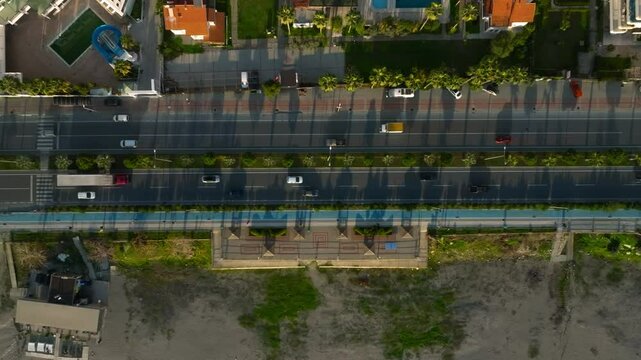 Drone view from above of a highway divided by palm trees with moving cars in a resort city, with residential buildings on one side and a beach on the other, in Mahmutlar, Alanya, Antalya, Turkey
