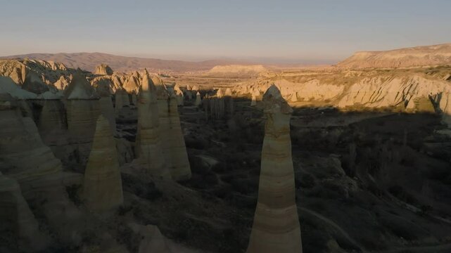 Drone flight between elongated unusual rock formations resembling chimneys or phalluses, illuminated by the sunset at the Love Valley in Cappadocia