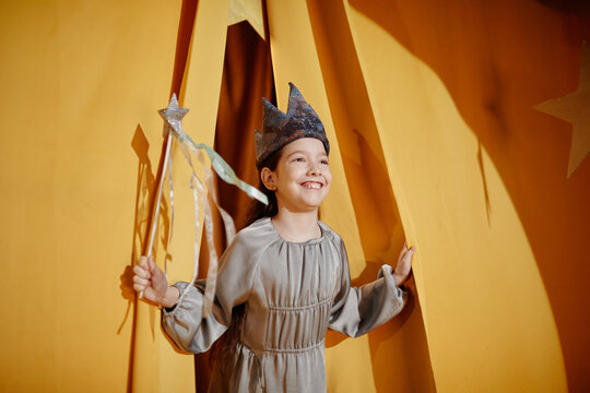 Smiling child wearing a crown and holding a magic wand, standing in a theater-like environment with yellow curtains, bringing light and joy to the scene through imagination and innocence