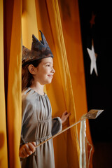 Young girl wearing a handmade blue crown smiling while peeking from behind decorative curtains. Background includes a star-shaped decoration hanging