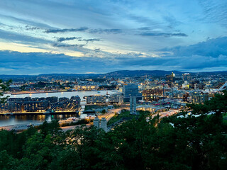Fototapeta premium Evening panorama of Oslo city viewed from Ekebergrestauranten, showcasing illuminated cityscape, roads, hills, and dramatic twilight sky in Norway.