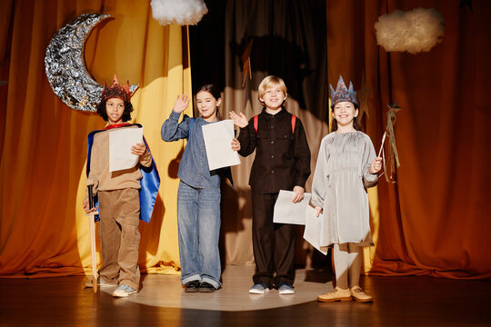 Four children on stage during a school play, holding scripts and wearing costumes, with golden curtains and stage props visible in background