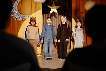 Group of children standing on stage during school play, wearing various costumes. Background...