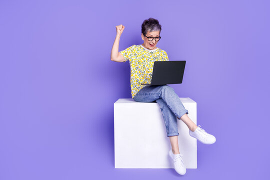 Happy elderly woman using a laptop, expressing excitement against purple background