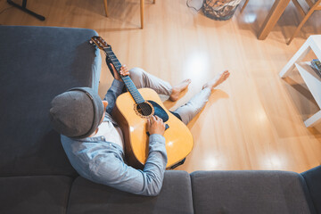Musician sits on a gray couch, casually strumming an acoustic guitar, immersed in a moment of...