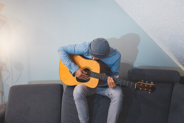 Musician sits on a gray couch, fully absorbed in playing an acoustic guitar, his posture reflecting...