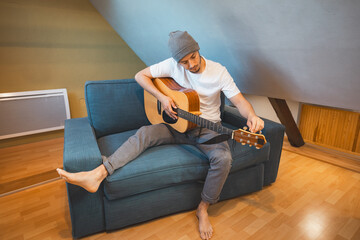 Musician 26 years old sits barefoot on a cozy blue couch in an attic room, strumming an acoustic...
