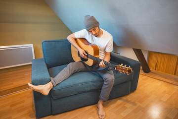 Musician 26 years old sits barefoot on a cozy blue couch in an attic room, strumming an acoustic...