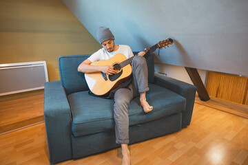 Musician 26 years old sits barefoot on a cozy blue couch in an attic room, strumming an acoustic...