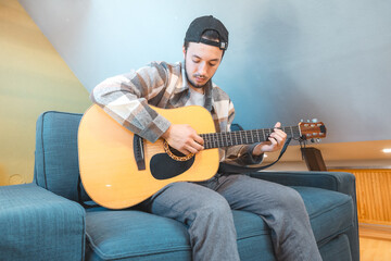 Young man with a relaxed demeanor sits barefoot on a cozy blue couch, strumming an acoustic guitar....