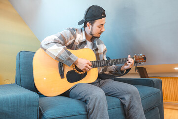 Young man with a relaxed demeanor sits barefoot on a cozy blue couch, strumming an acoustic guitar....