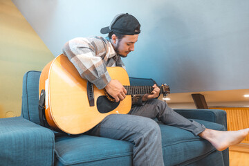 Young man with a relaxed demeanor sits barefoot on a cozy blue couch, strumming an acoustic guitar....