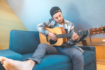 Young man with a relaxed demeanor sits barefoot on a cozy blue couch, strumming an acoustic guitar....